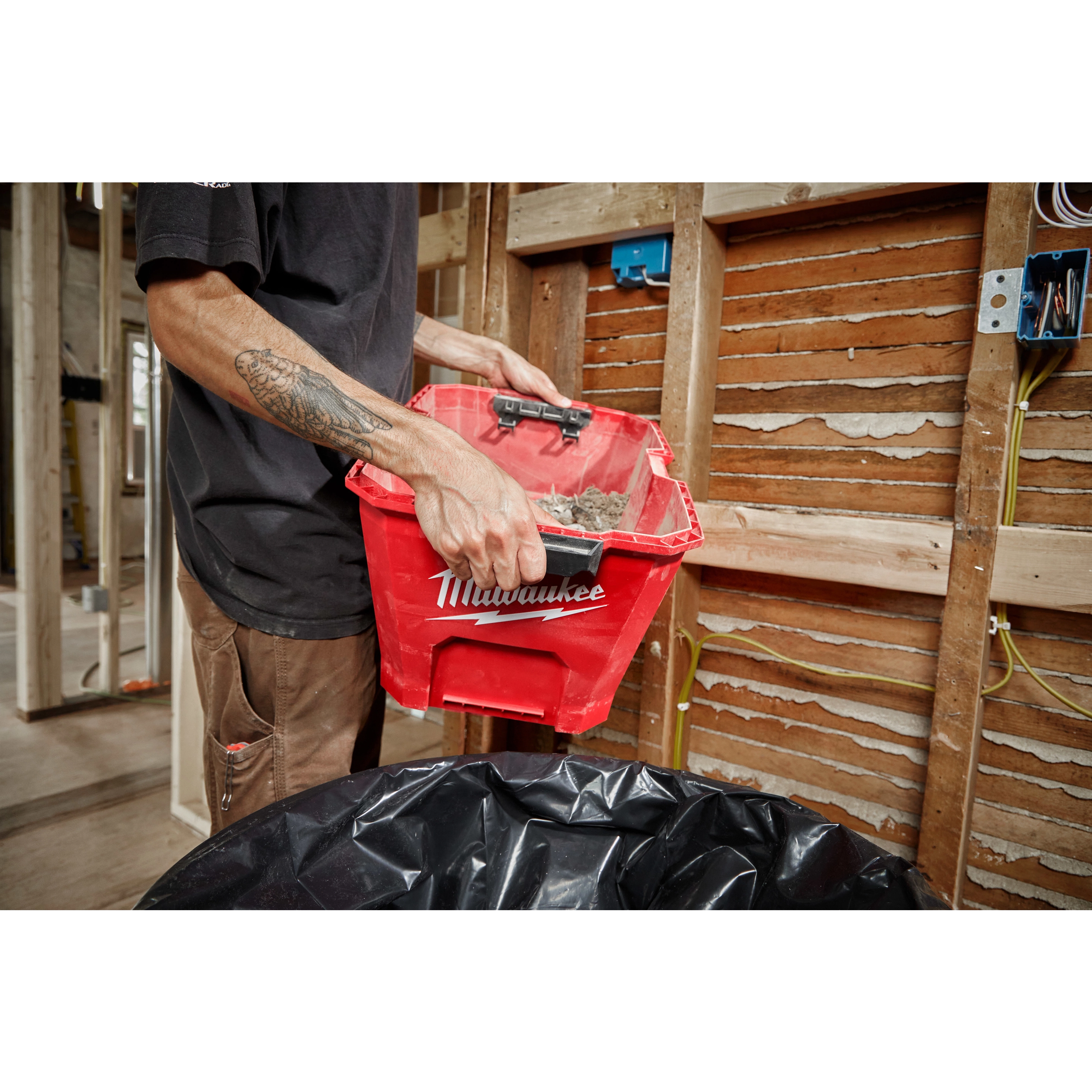 A person empties debris from a Milwaukee 6 Gallon Wet/Dry Vacuum Tank into a black garbage bag at a construction site.