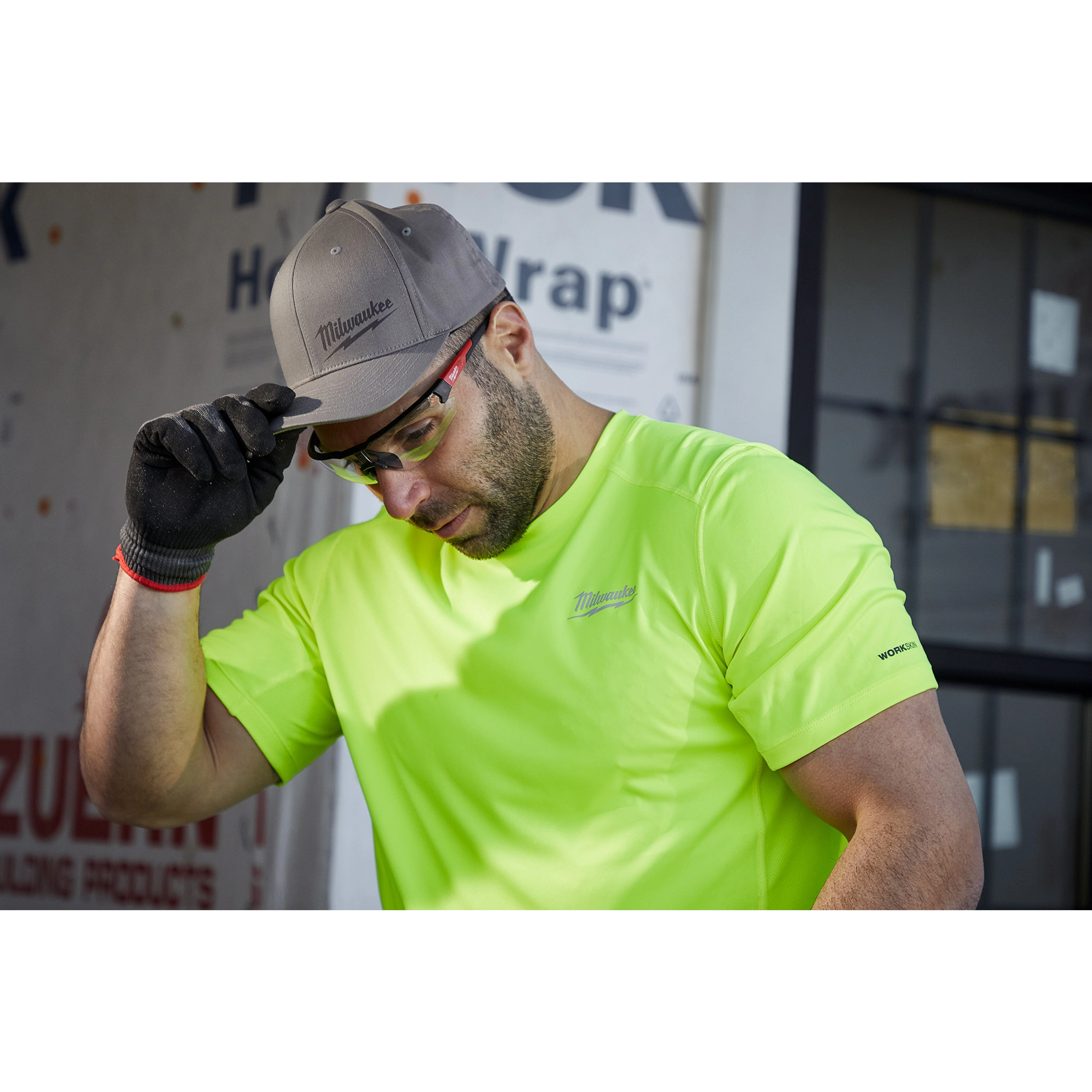 Man wearing a Milwaukee Fitted Hat and neon green Milwaukee work shirt, lifting the hat's brim, standing in front of a building.