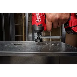 A hand uses a red power drill with a 1-3/8" Shockwave Impact Hole Saw to cut through a piece of metal.