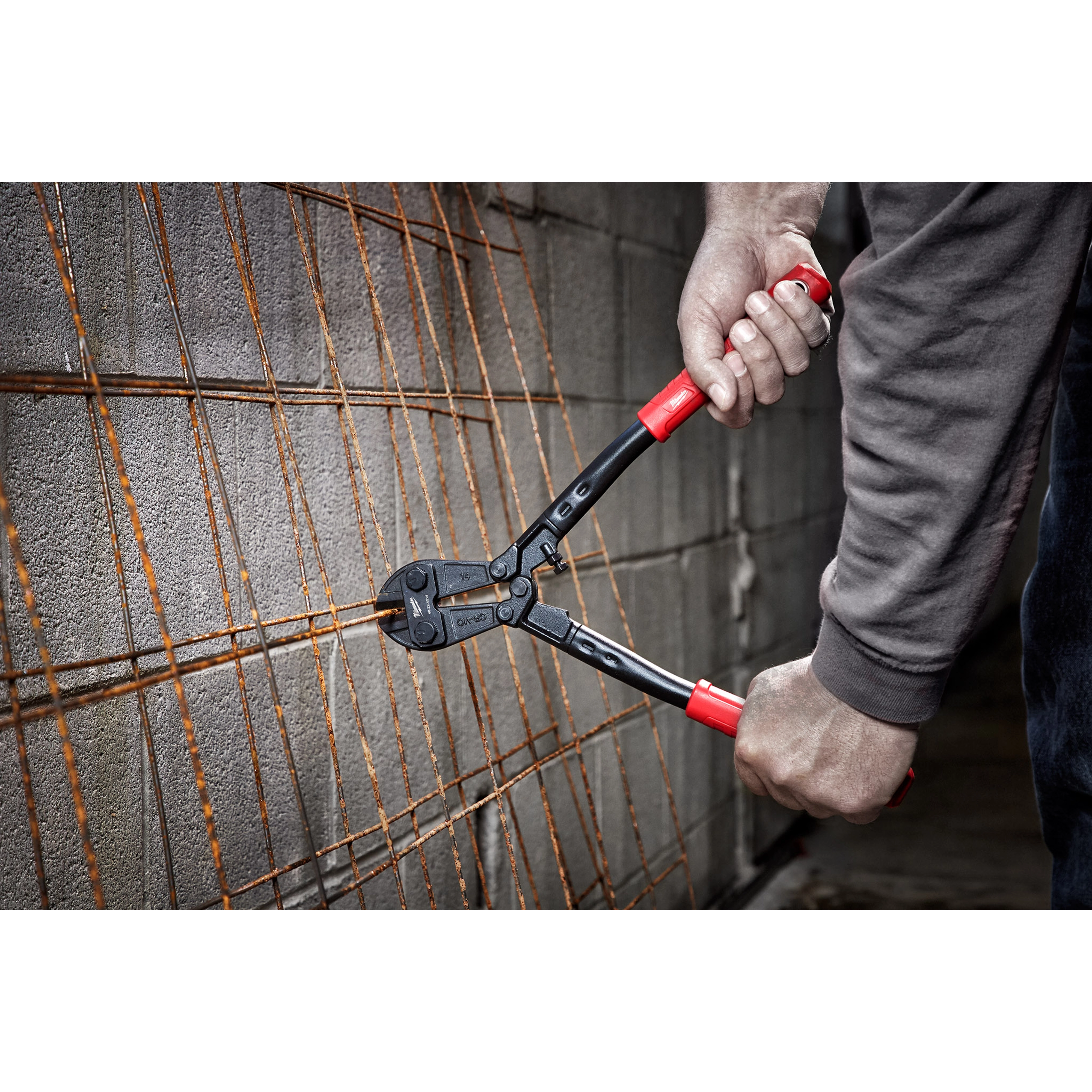 Hands using a 14" Bolt Cutter with red handles to cut rusted metal wire mesh against a concrete block wall backdrop.