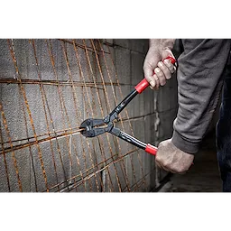 Hands using a 14" Bolt Cutter with red handles to cut rusted metal wire mesh against a concrete block wall backdrop.