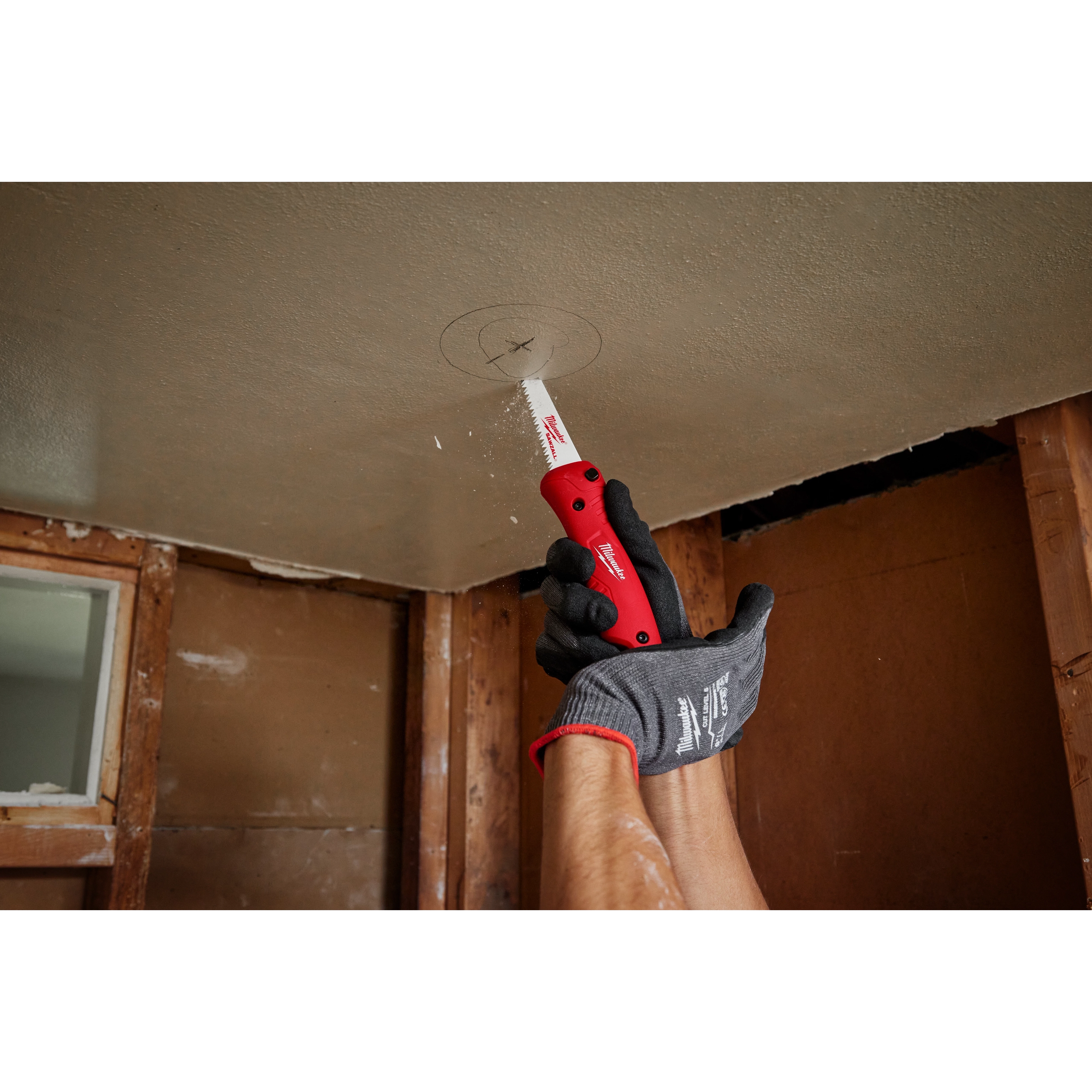 A person in gloves uses a Folding Jab Saw to cut a marked circle in a ceiling during a renovation project.