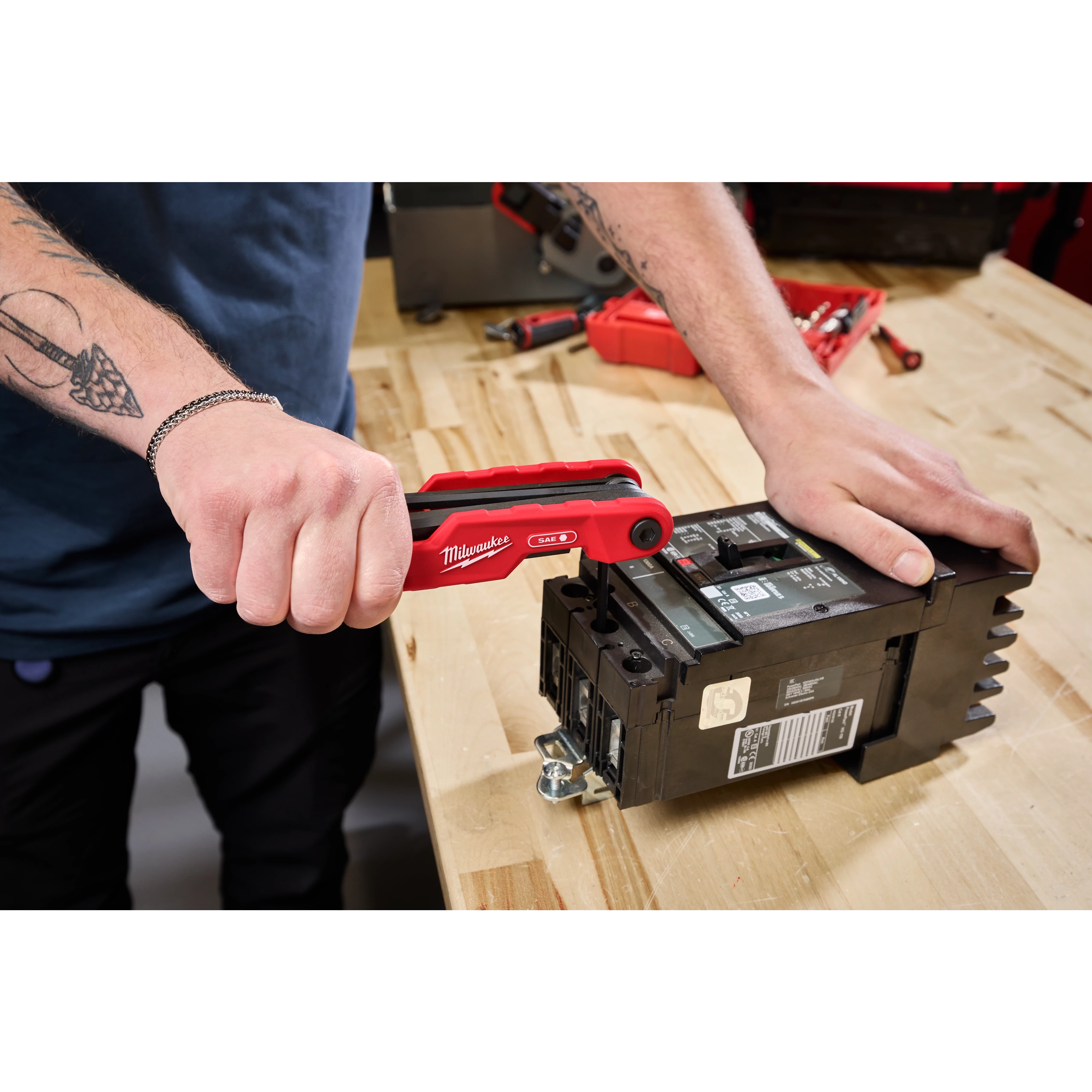 A person uses a Milwaukee hand tool to work on an electrical component on a wooden workbench. The person has tattoos on both arms and wears a chain bracelet on their right wrist.