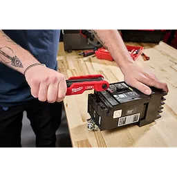 A person uses a Milwaukee hand tool to work on an electrical component on a wooden workbench. The person has tattoos on both arms and wears a chain bracelet on their right wrist.