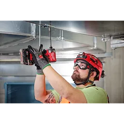 A construction worker wearing Vented Low-Profile Goggles - Clear Dual Coat Lens uses a red cordless drill to work on overhead metal ducting.