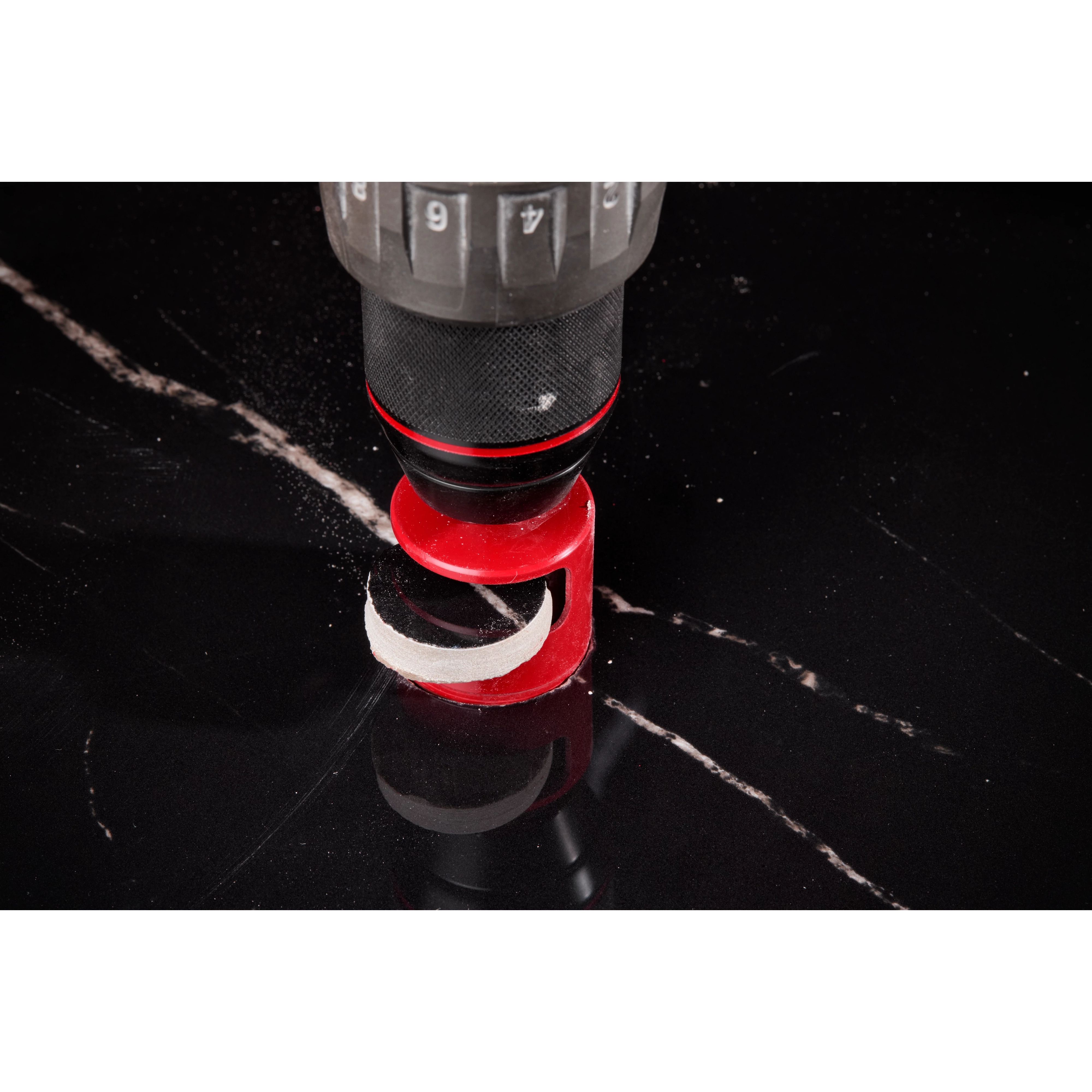 A close-up of a drill with a Milwaukee® 1-3/8" DIAMOND MAX™ Hole Saw Bit cutting into a black, glossy surface with white veining. The red hole saw bit is mounted on the drill, which is in the process of making a circular cut.