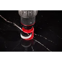A close-up of a drill with a Milwaukee® 1-3/8" DIAMOND MAX™ Hole Saw Bit cutting into a black, glossy surface with white veining. The red hole saw bit is mounted on the drill, which is in the process of making a circular cut.