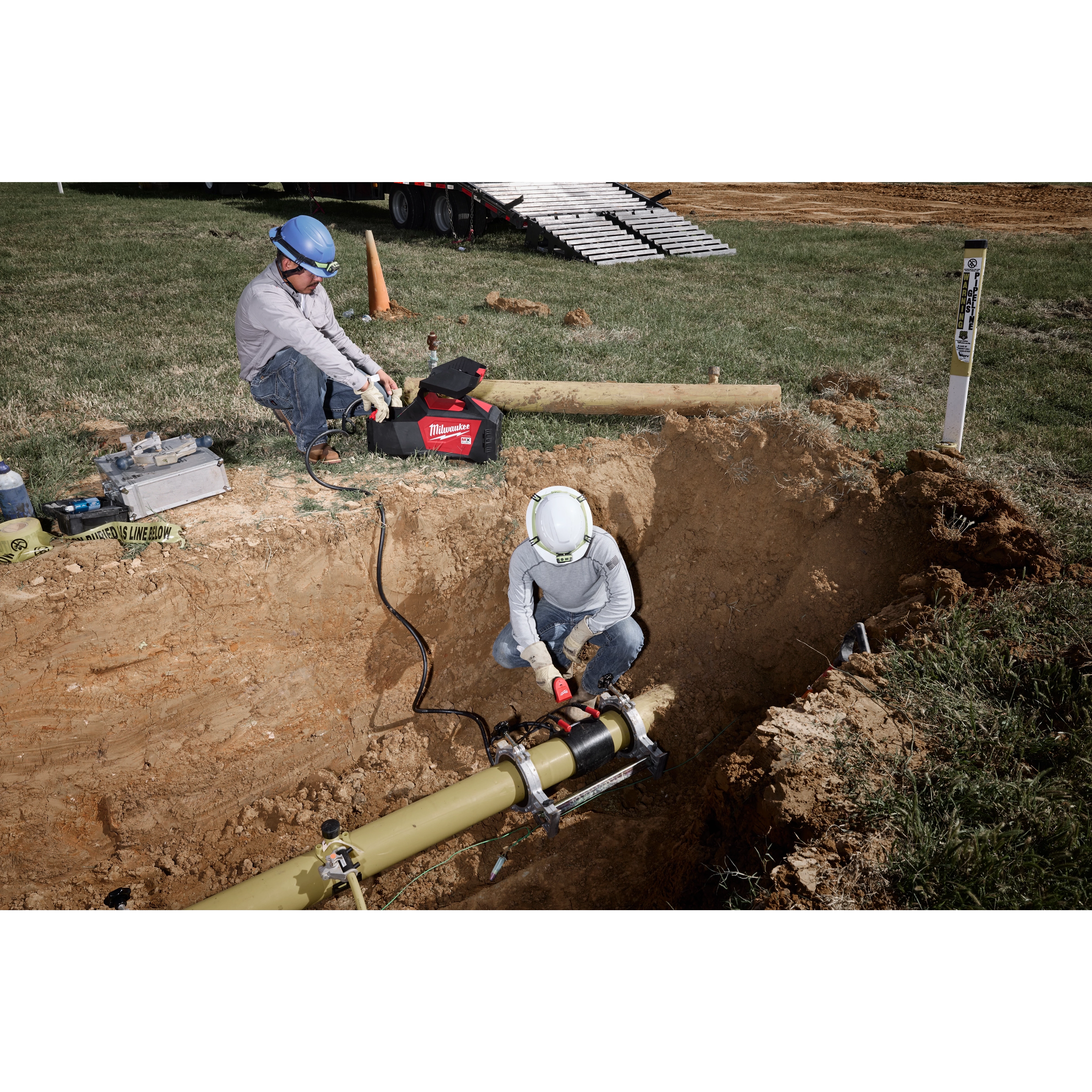 Two workers in safety gear are using the MX FUEL™ Electrofusion Processor on a yellow pipe in a dug-out area. One worker operates the processor while the other monitors on the surface. Equipment and safety cones are visible nearby.