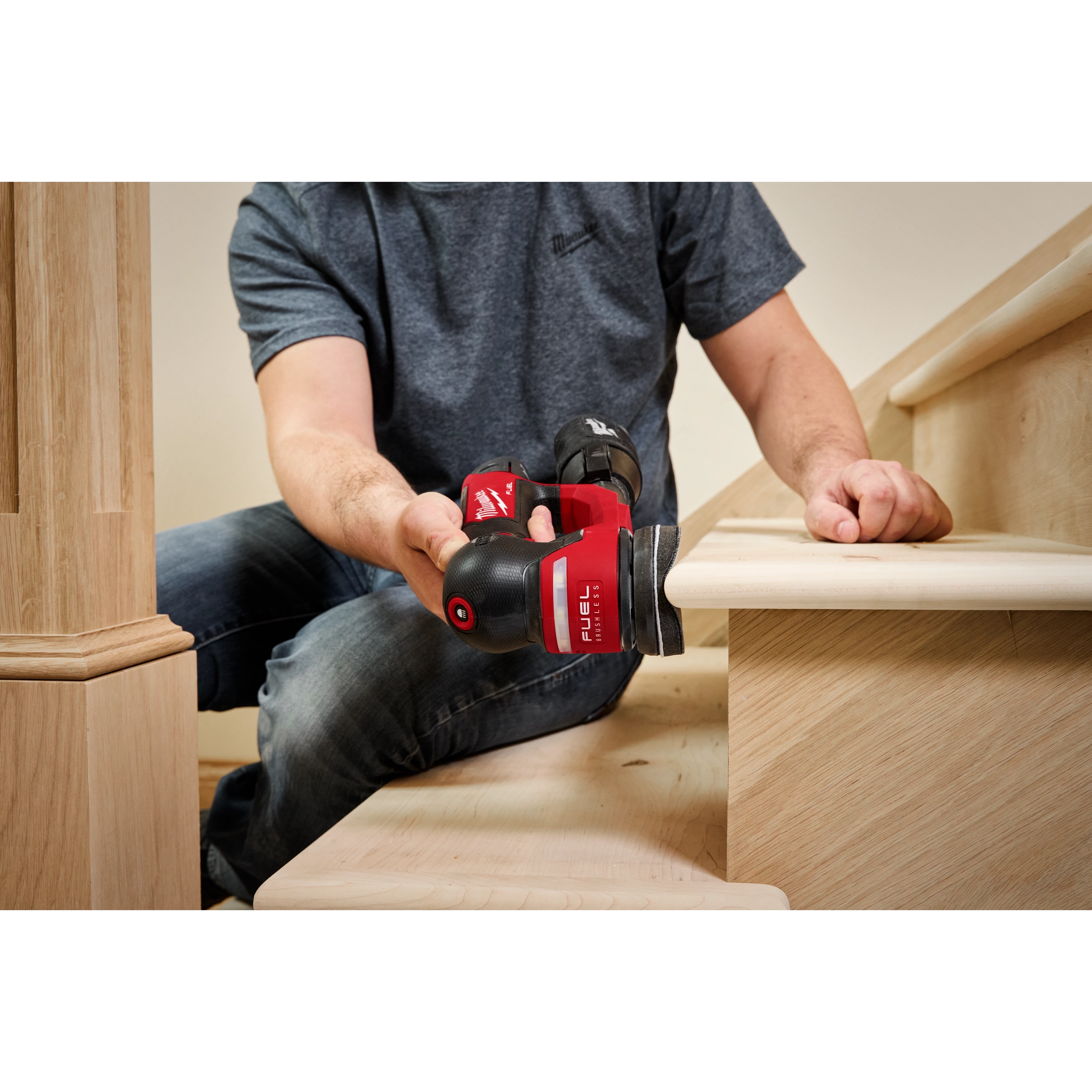 A person is using an M12 FUEL™ 3" Random Orbital Detail Sander to sand the edge of a wooden stair tread. The person is wearing a gray shirt and jeans, focusing on the task. The sander is red and black.