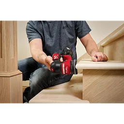 A person is using an M12 FUEL™ 3" Random Orbital Detail Sander to sand the edge of a wooden stair tread. The person is wearing a gray shirt and jeans, focusing on the task. The sander is red and black.