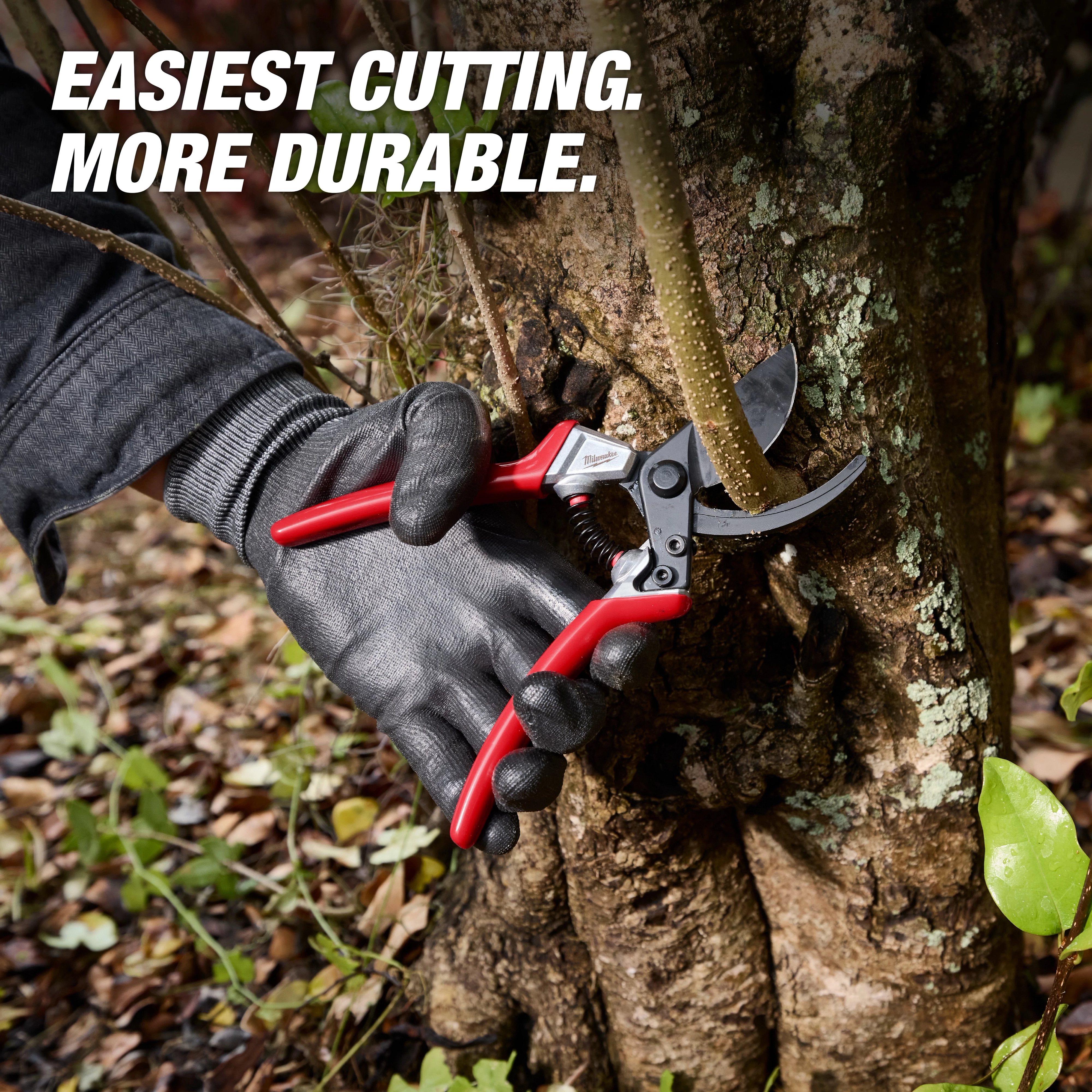 A gloved hand uses a 1" Pruner with red handles to cut a branch against a tree trunk. The image features the text "EASIEST CUTTING. MORE DURABLE." surrounded by leaves and outdoor ground cover.