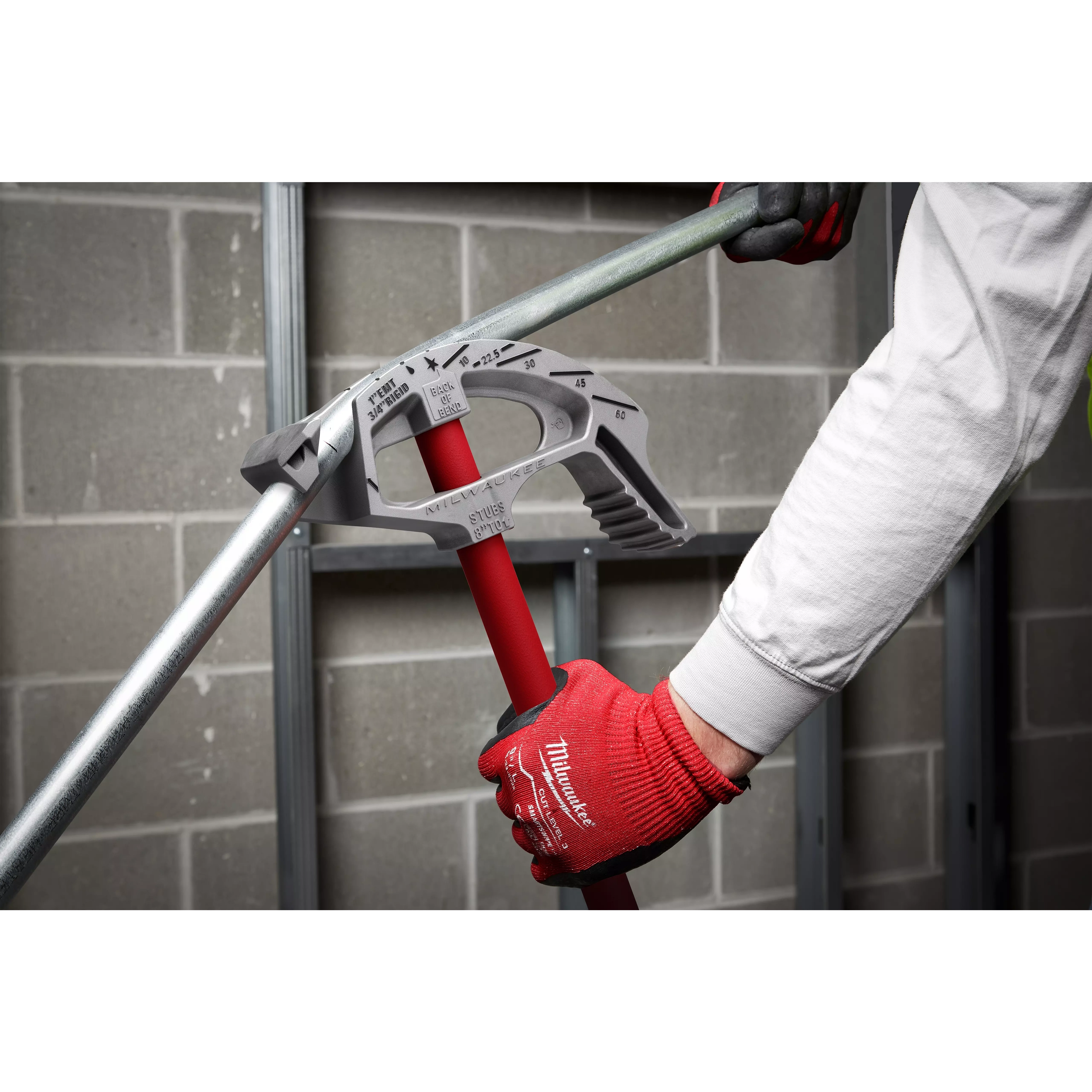 A person wearing a white shirt and red gloves uses Offset Jobsite Scissors to cut a metal pipe against a concrete block wall background.