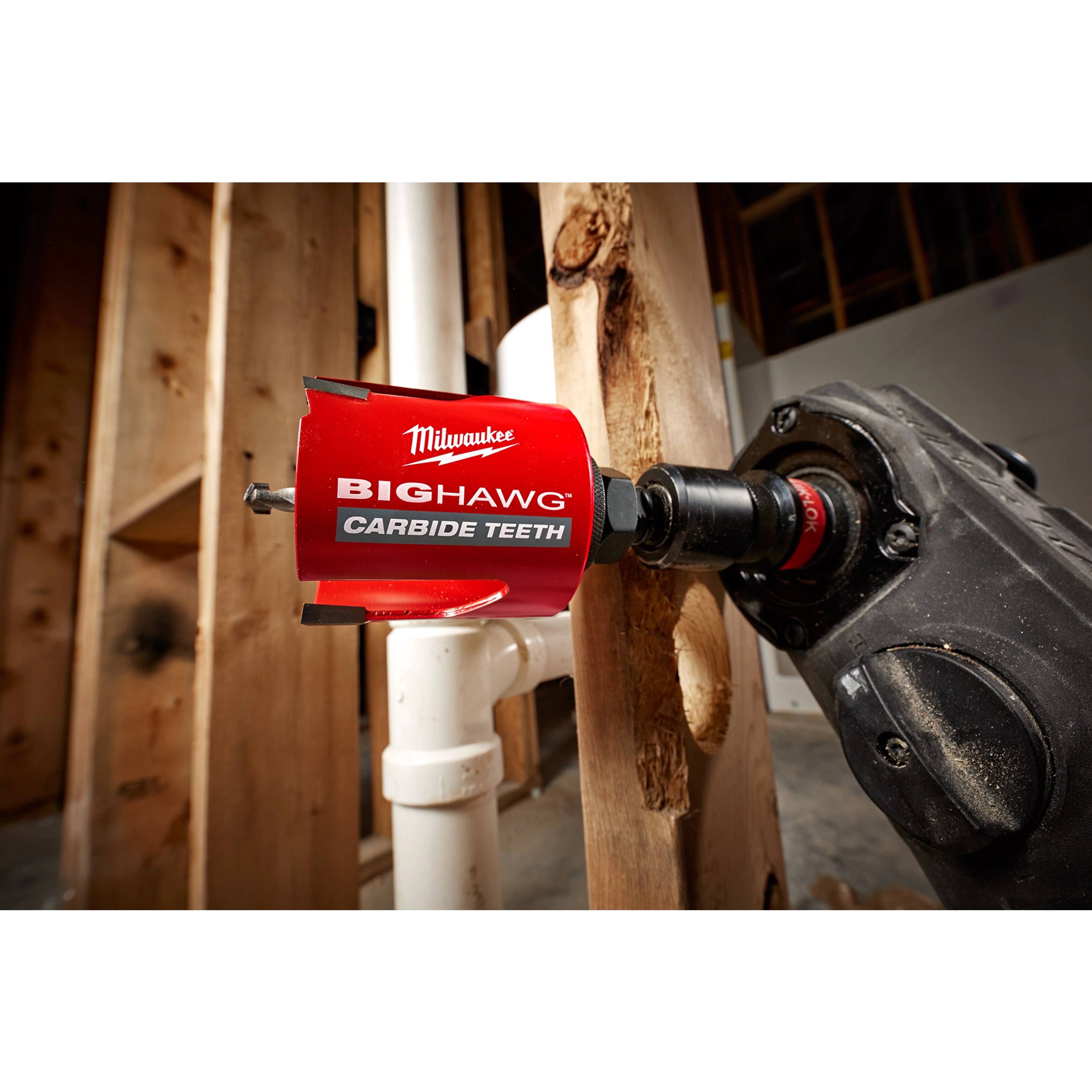 Red 2-9/16" Big Hawg with Carbide Teeth hole saw attached to a black drill, cutting through a wooden beam next to PVC pipes.