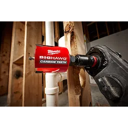 Red 2-9/16" Big Hawg with Carbide Teeth hole saw attached to a black drill, cutting through a wooden beam next to PVC pipes.