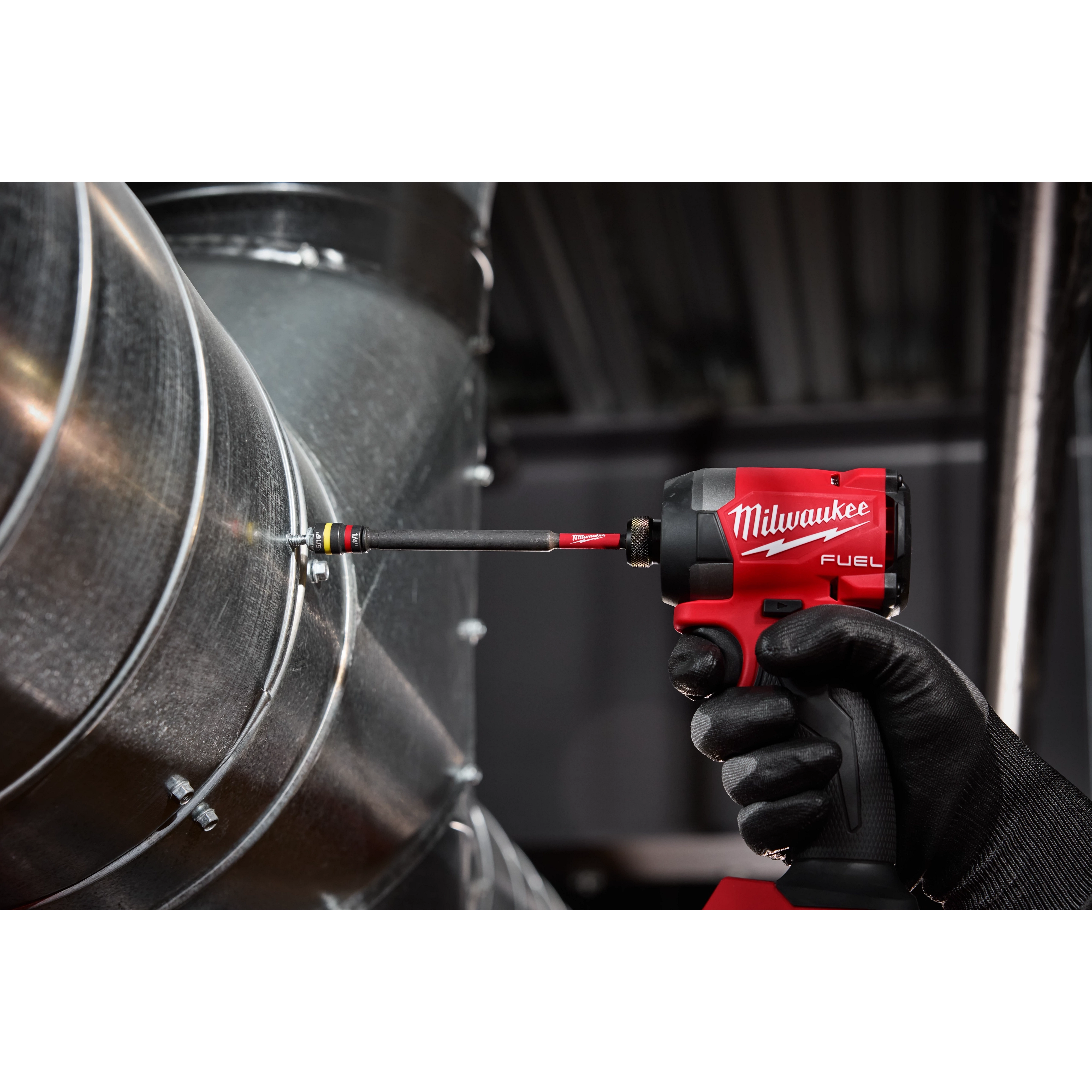 The SHOCKWAVE™ Impact Duty 1/4” and 5/16” x 6” QUIK-CLEAR™ 2-in-1 Magnetic Nut Driver is being used on a metallic duct. The tool is attached to a red Milwaukee power drill, and a gloved hand is holding it while tightening a bolt.