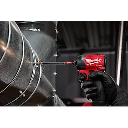 The SHOCKWAVE™ Impact Duty 1/4” and 5/16” x 6” QUIK-CLEAR™ 2-in-1 Magnetic Nut Driver is being used on a metallic duct. The tool is attached to a red Milwaukee power drill, and a gloved hand is holding it while tightening a bolt.