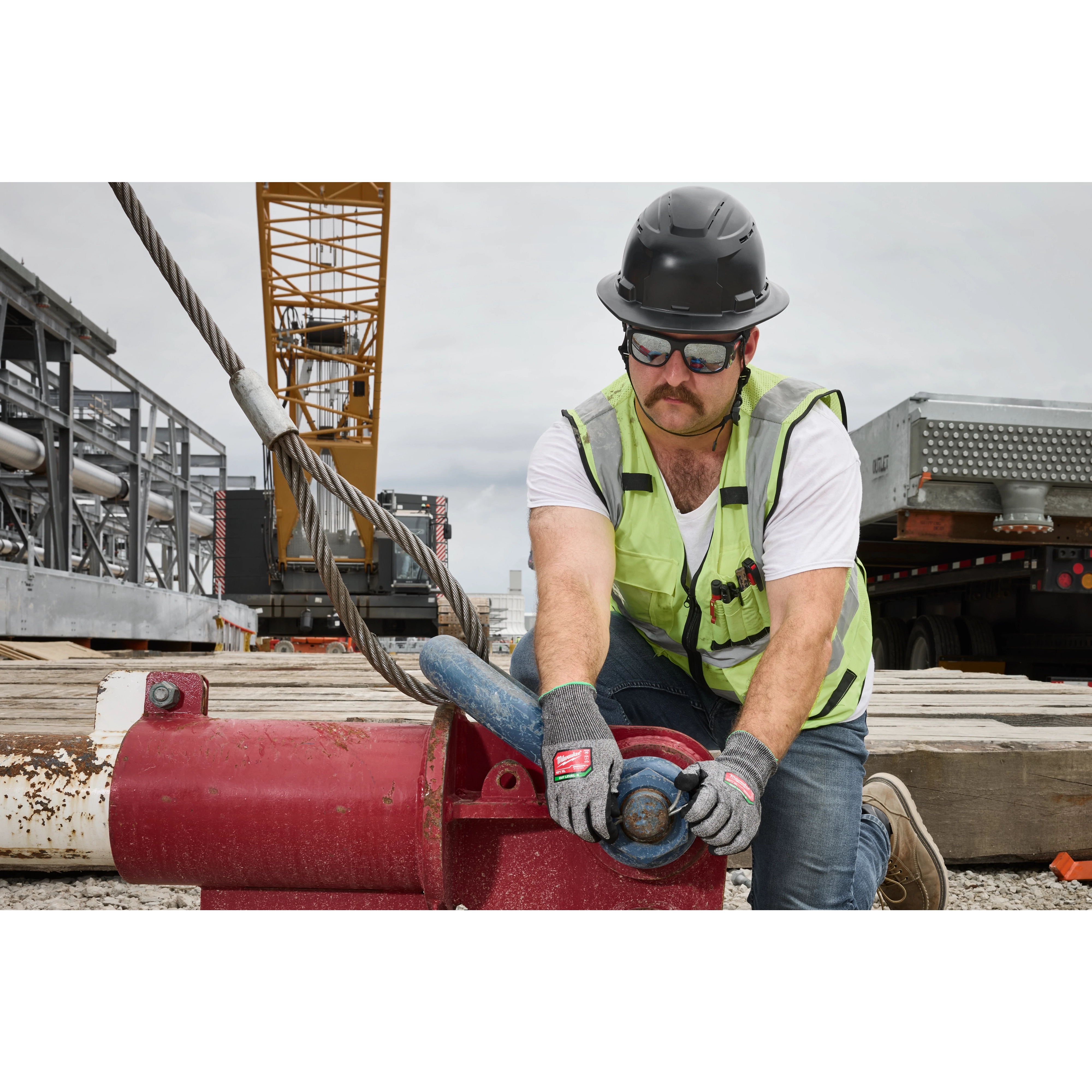 A construction worker wearing a hard hat, neon safety vest, and Full Frame Safety Glasses with Removable Side Shields – Silver Mirrored Anti-Scratch Lenses is kneeling while operating industrial equipment at a worksite featuring scaffolding and a crane.