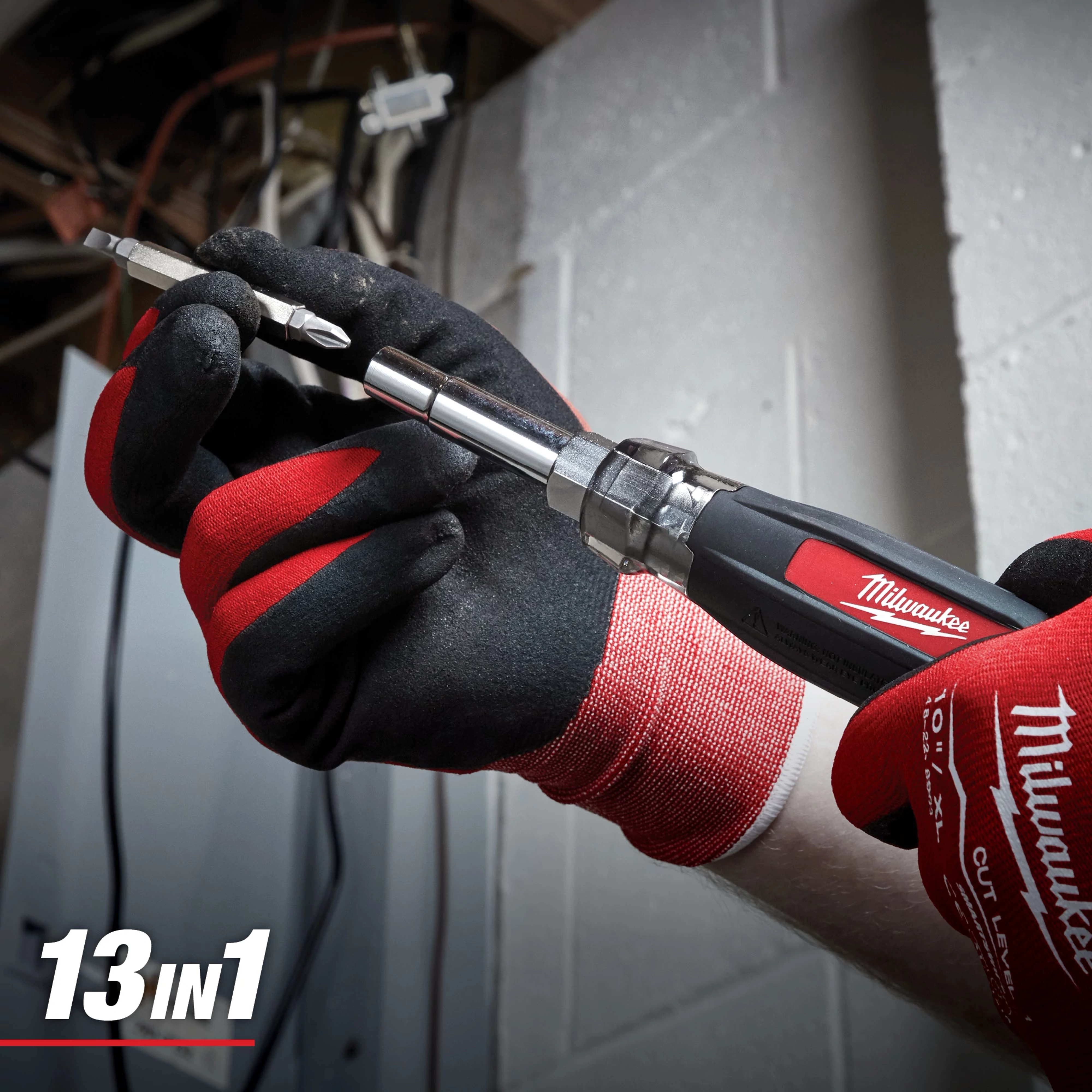 A person wearing Milwaukee-branded work gloves holds a Milwaukee 13-in-1 Multi-Bit Screwdriver. The screwdriver has multiple interchangeable bits and a textured black and red handle for grip.