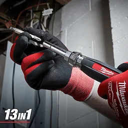 A person wearing Milwaukee-branded work gloves holds a Milwaukee 13-in-1 Multi-Bit Screwdriver. The screwdriver has multiple interchangeable bits and a textured black and red handle for grip.