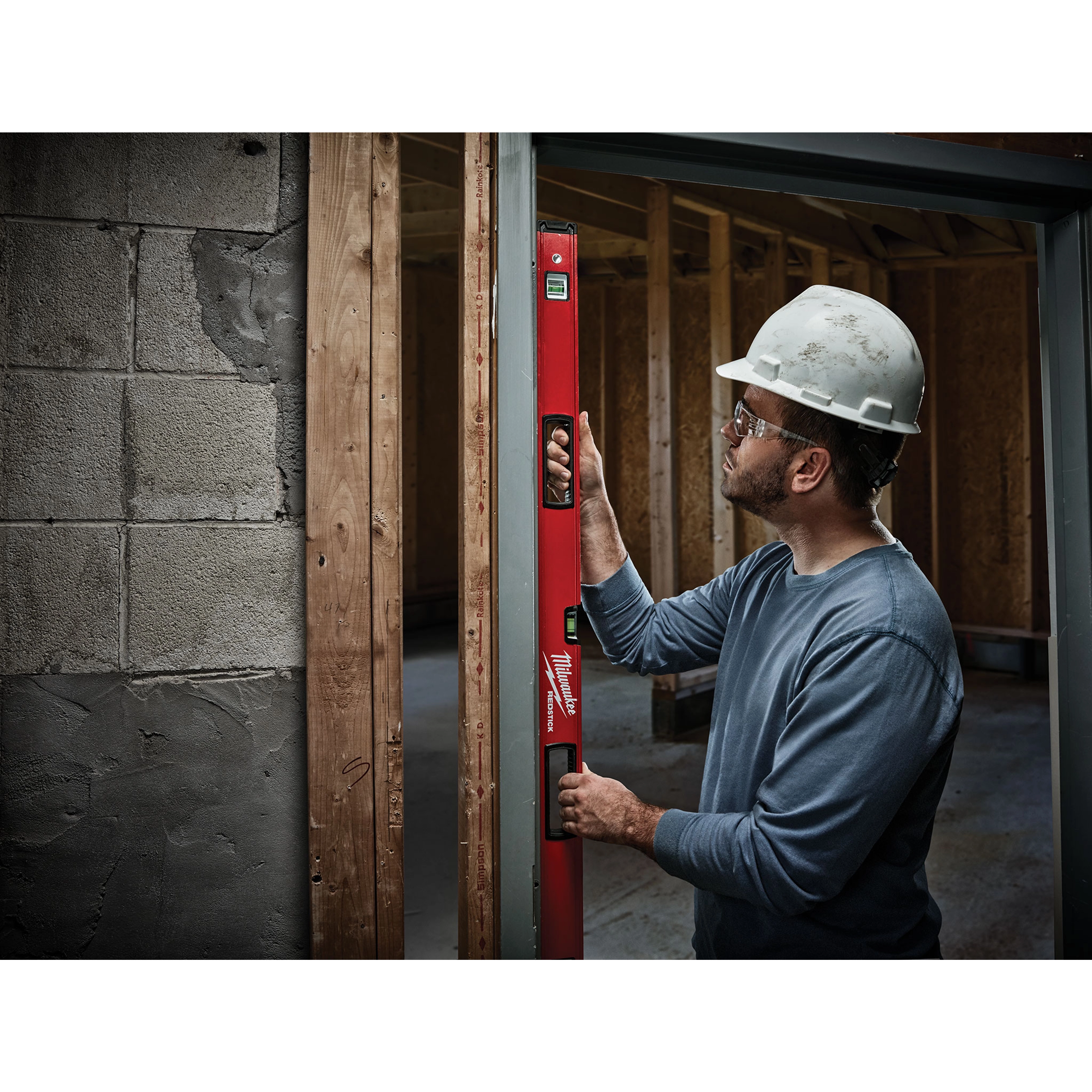 A construction worker using a REDSTICK Level to ensure vertical accuracy in a wooden frame structure at a building site.