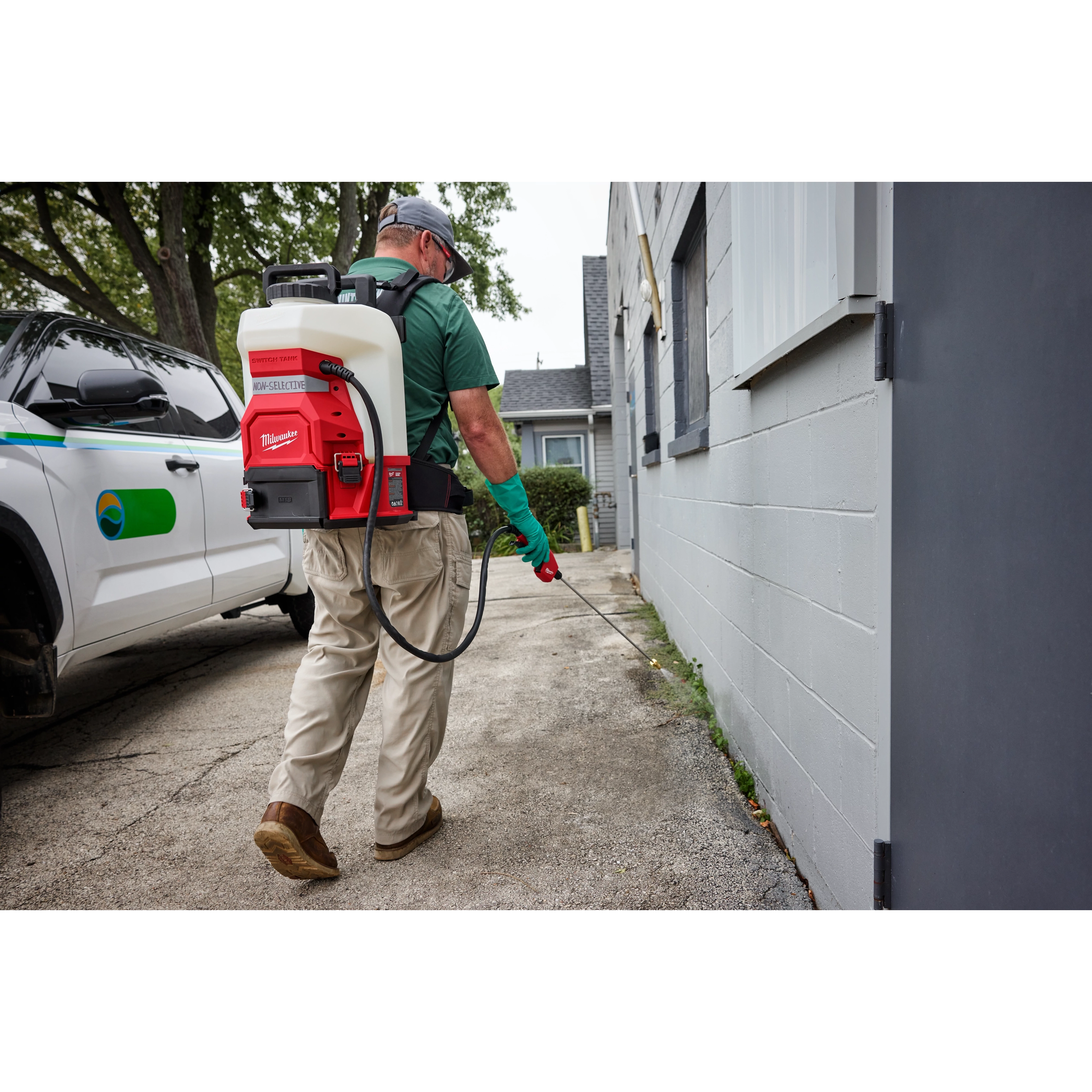 A person wearing gloves and a cap uses the M18™ 4-Gallon Backpack Sprayer w/ SWITCH TANK™ to spray along the side of a building. They are walking on a paved surface, and a work truck is parked nearby. The sprayer is white with red accents and a black frame.