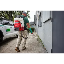A person wearing gloves and a cap uses the M18™ 4-Gallon Backpack Sprayer w/ SWITCH TANK™ to spray along the side of a building. They are walking on a paved surface, and a work truck is parked nearby. The sprayer is white with red accents and a black frame.