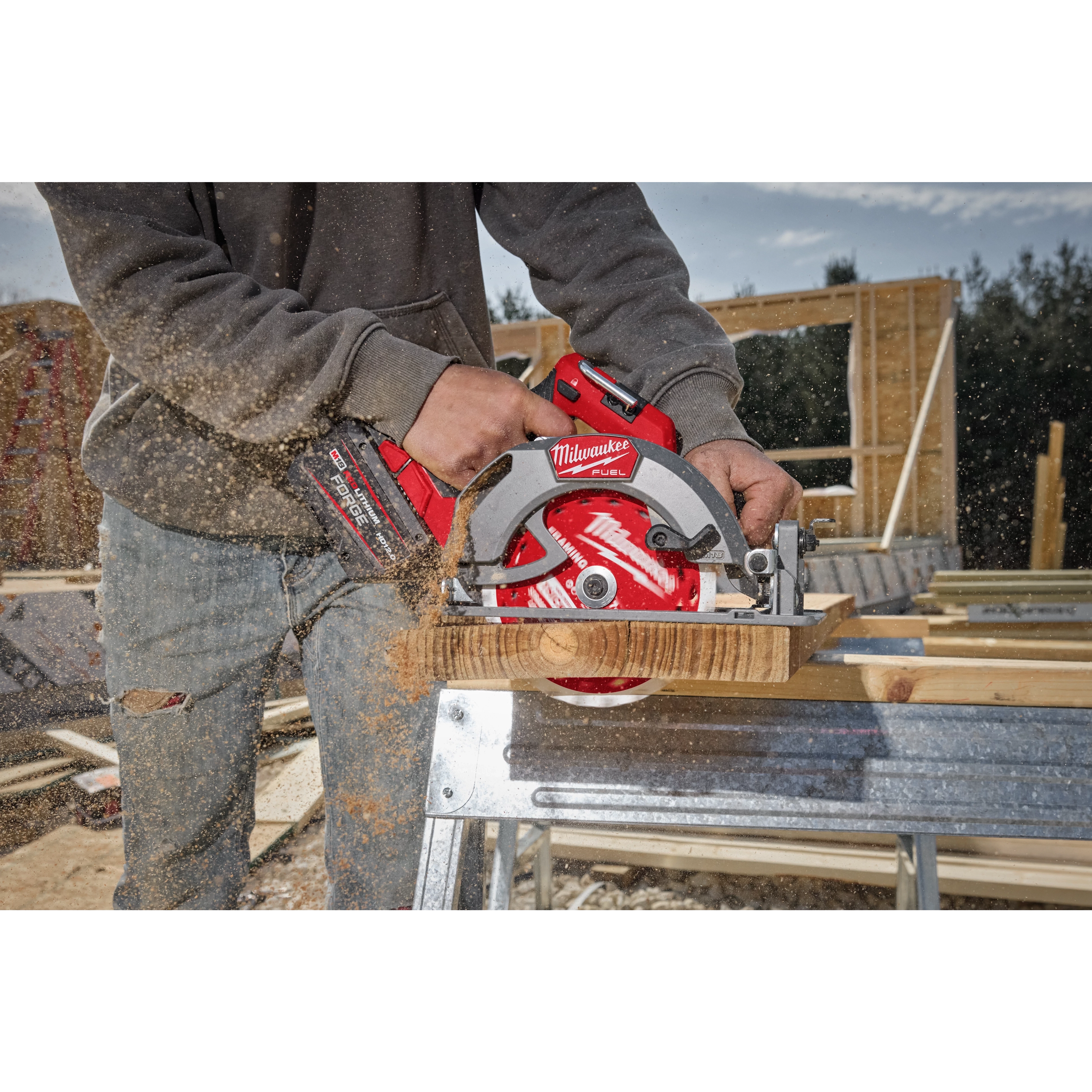 A person operates an M18 FUEL™ 7-1/4” Circular Saw to cut through a piece of wood. The saw is red and black, producing wood dust as it cuts. The scene appears to be on a construction site with wooden structures in the background.