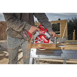 A person operates an M18 FUEL™ 7-1/4” Circular Saw to cut through a piece of wood. The saw is red and black, producing wood dust as it cuts. The scene appears to be on a construction site with wooden structures in the background.