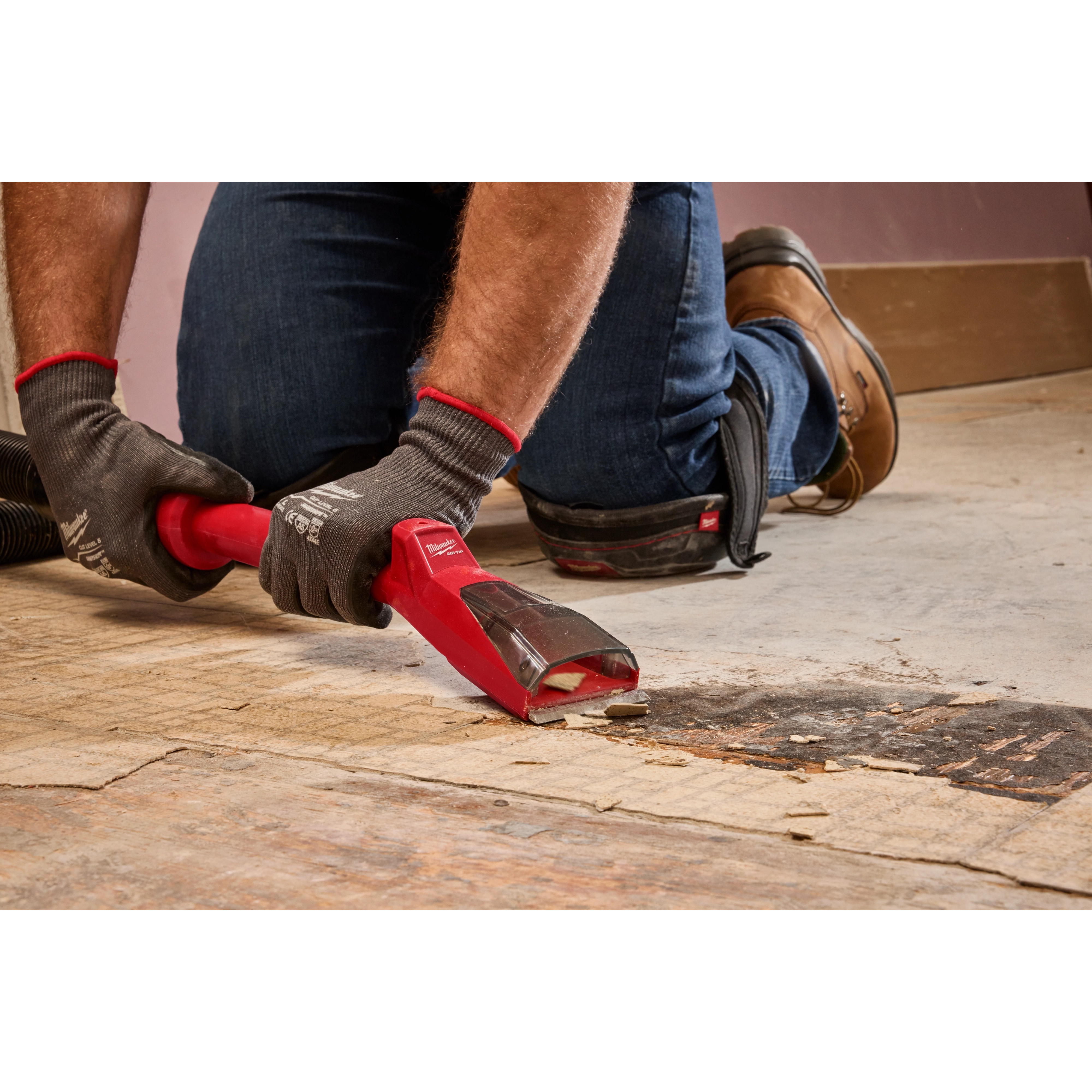 Person using the AIR-TIP Debris Scraper to remove debris from a floor while wearing work gloves, jeans, and brown boots.