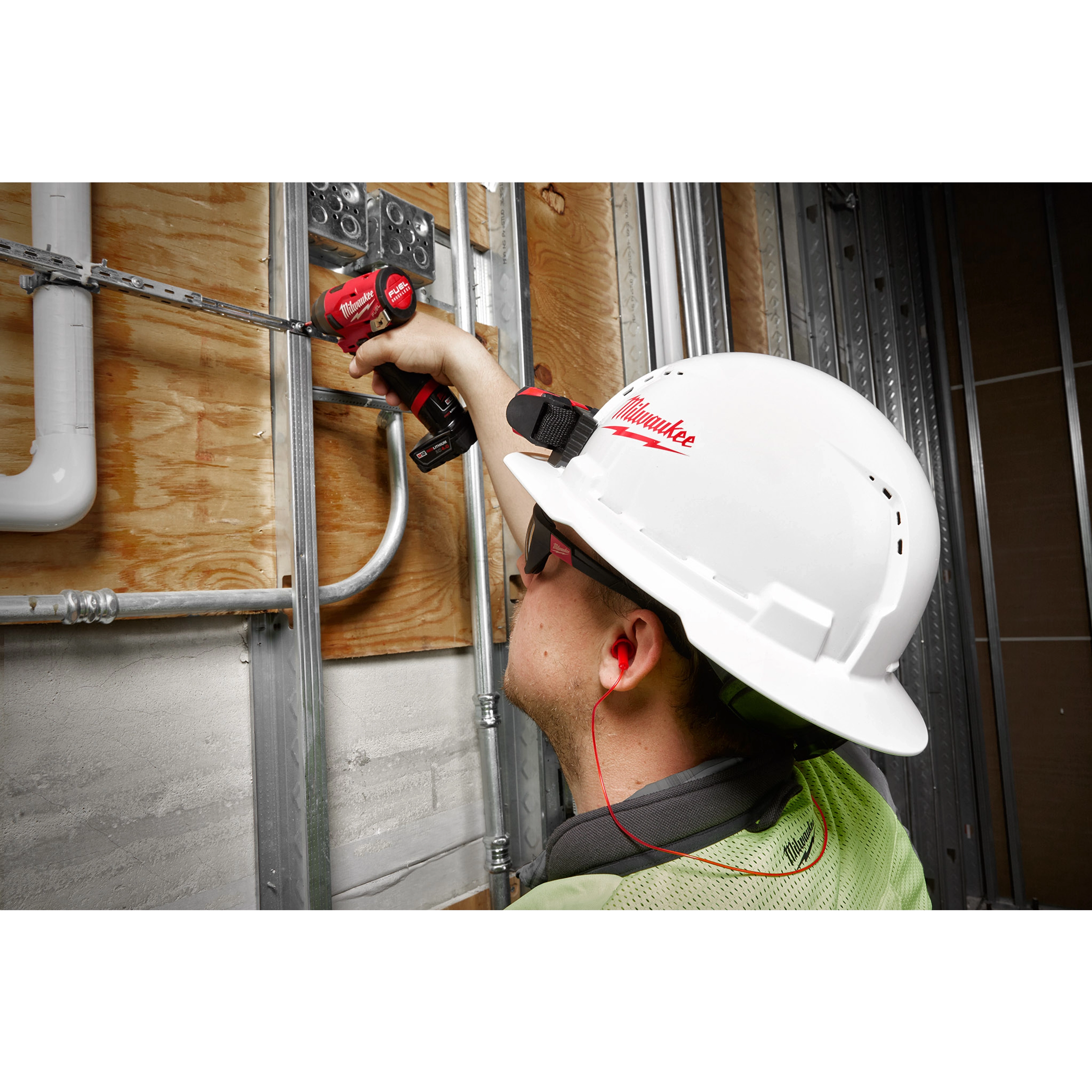 Worker wearing a white hard hat and safety glasses uses a red power drill while wearing red Corded Ear Plugs on a construction site.