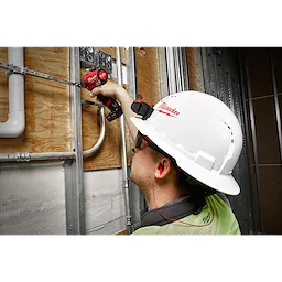 Worker wearing a white hard hat and safety glasses uses a red power drill while wearing red Corded Ear Plugs on a construction site.
