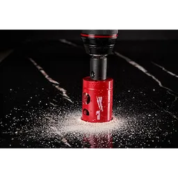 A Milwaukee® 1-3/8" DIAMOND MAX™ Hole Saw is cutting through a dark, polished surface, producing a ring of light-colored dust. The red hole saw is attached to a black drill, showcasing its diamond-enforced teeth designed for durability.