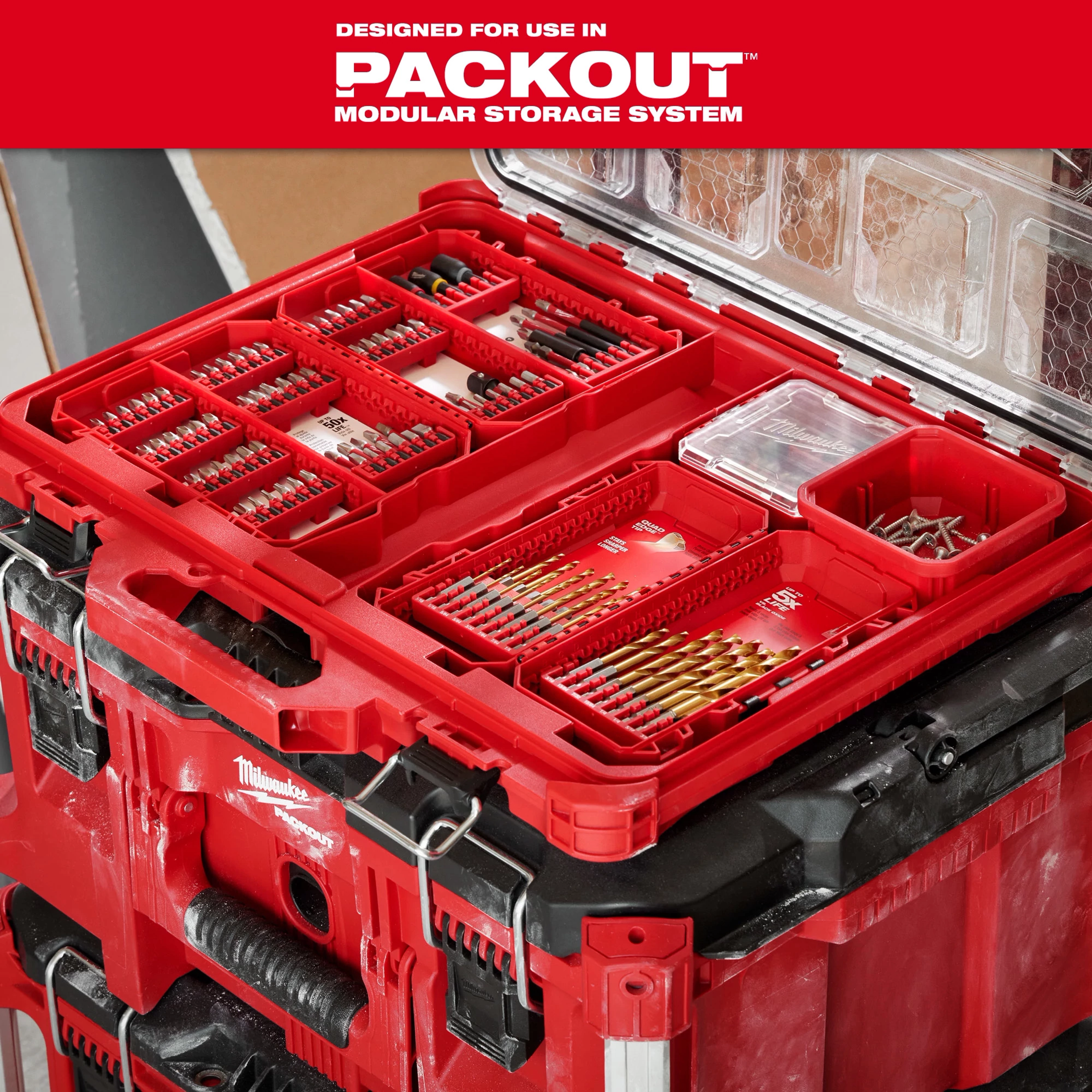 The image features a red storage system designed for use in PACKOUT Modular Storage System. It contains organized compartments with various drill bits, screws, and fasteners. The case sits atop another PACKOUT unit and includes clear plastic lids for visibility and protection of contents.