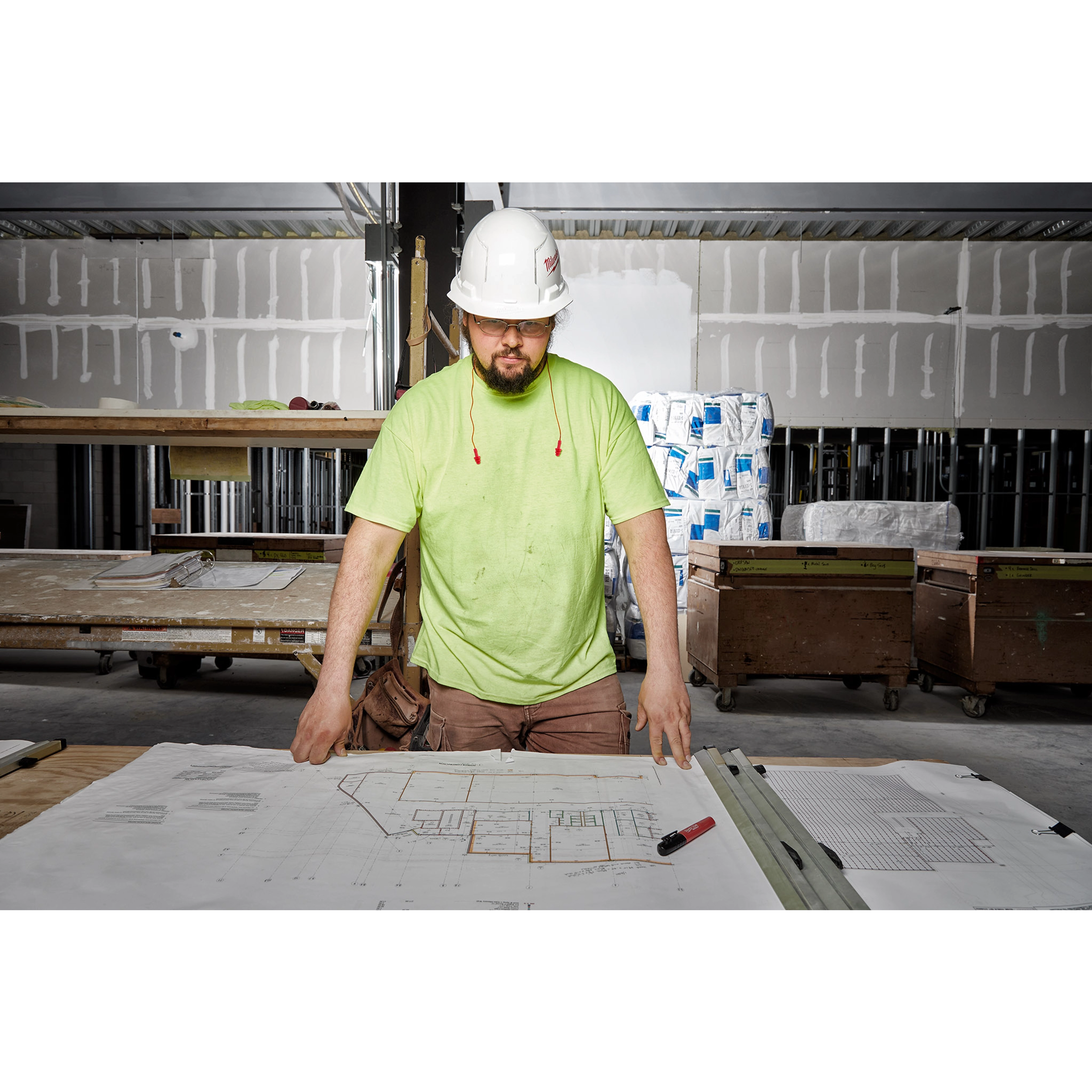 Construction worker wearing Ear%20Plugs stands over blueprints in a partially completed building, wearing a neon green shirt and white hard hat.