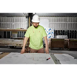 Construction worker wearing Ear%20Plugs stands over blueprints in a partially completed building, wearing a neon green shirt and white hard hat.