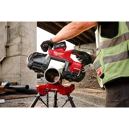 A person wearing gloves and a safety vest operates the M18 FUEL™ Deep Cut Dual Trigger Band Saw w/ ONE-KEY™ to cut a pipe on a construction site. The tool features a red and black color scheme and is mounted on a stand.