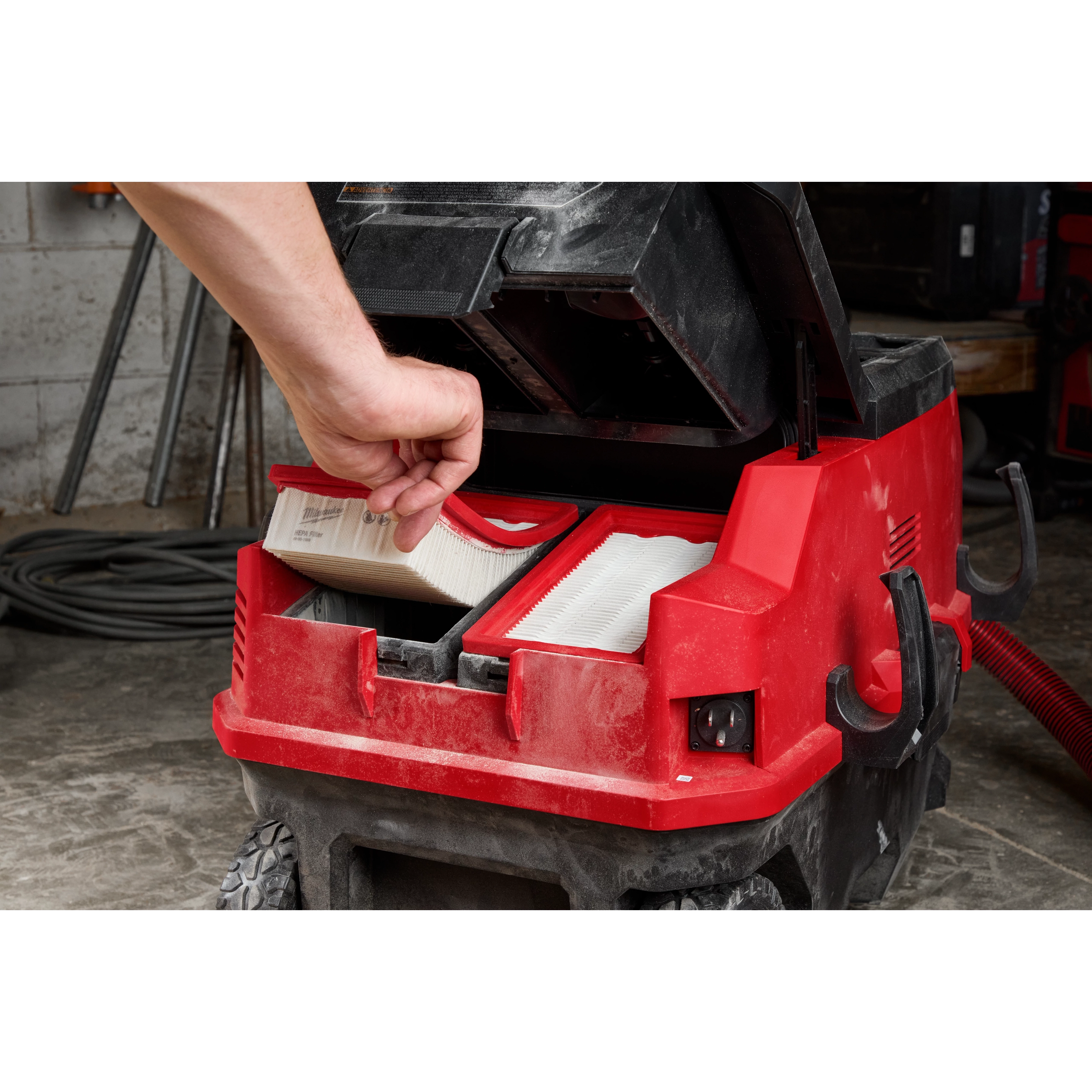 A hand is seen inserting a HEPA filter into a red vacuum cleaner. Another HEPA filter is already in place. The vacuum cleaner is open and located in a workshop setting with tools and coiled cords in the background. HEPA Filter (2PK) is indicated.