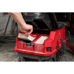 A hand is seen inserting a HEPA filter into a red vacuum cleaner. Another HEPA filter is already in place. The vacuum cleaner is open and located in a workshop setting with tools and coiled cords in the background. HEPA Filter (2PK) is indicated.