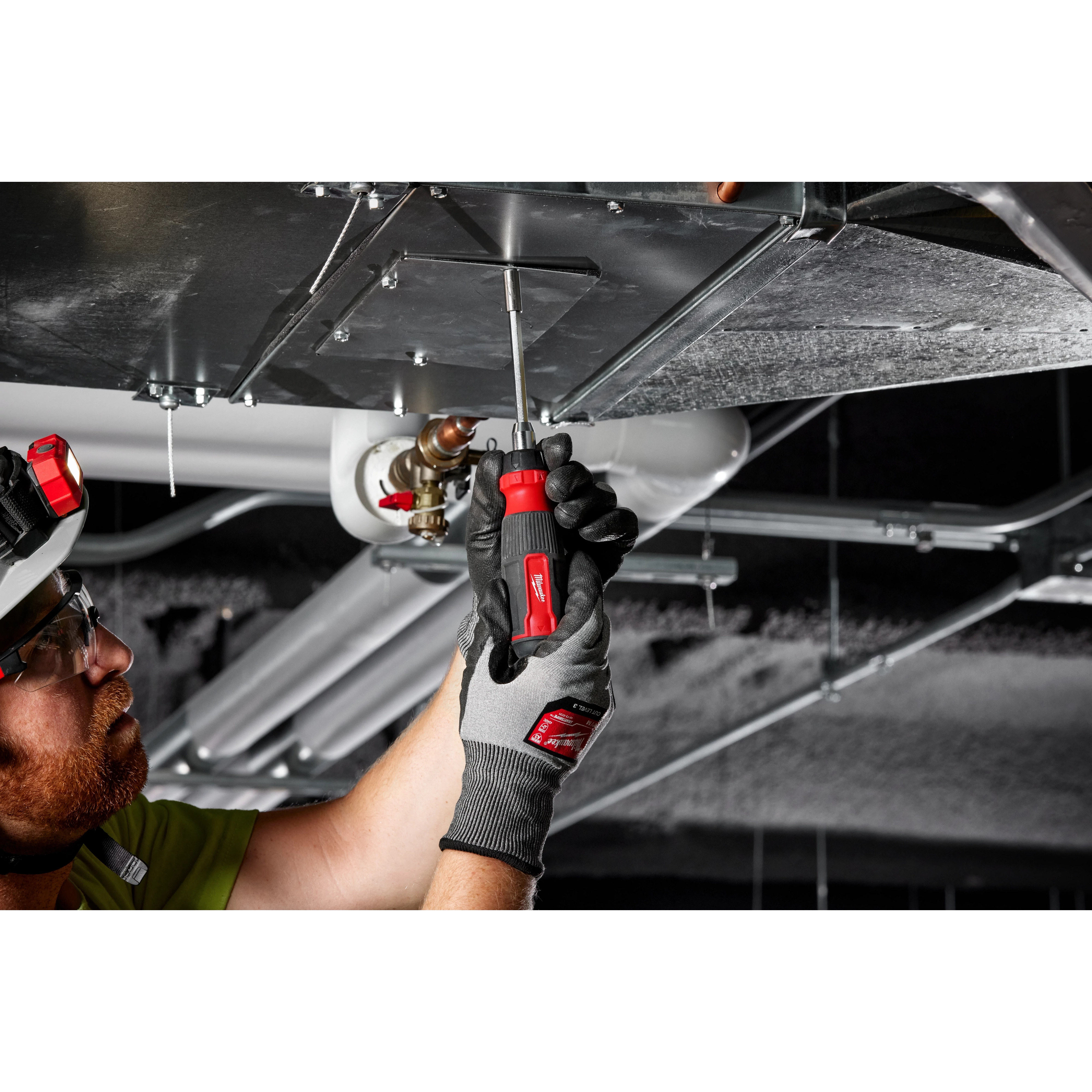 A person is using a 14-in-1 Ratcheting Multi-Bit Screwdriver to tighten a metal panel on a duct system in an industrial setting.
