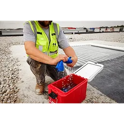 A construction worker kneels and wrings out a blue Cooling PVA Towel over a red cooler on a gravel-covered rooftop. The worker is wearing a neon yellow safety vest and grey shirt, with a cityscape visible in the background.