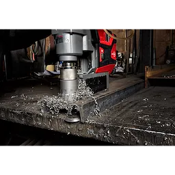 Annular Cutter attached to a red and gray power drill, drilling into metal with metal shavings scattered on the surface.