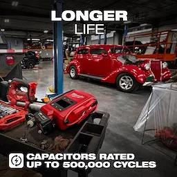 The M18™ HOTSHOT™ Jump Starter is on a table in a garage with tools and a classic red car in the background. The image highlights the product's durability with text "LONGER LIFE" and "CAPACITORS RATED UP TO 500,000 CYCLES."