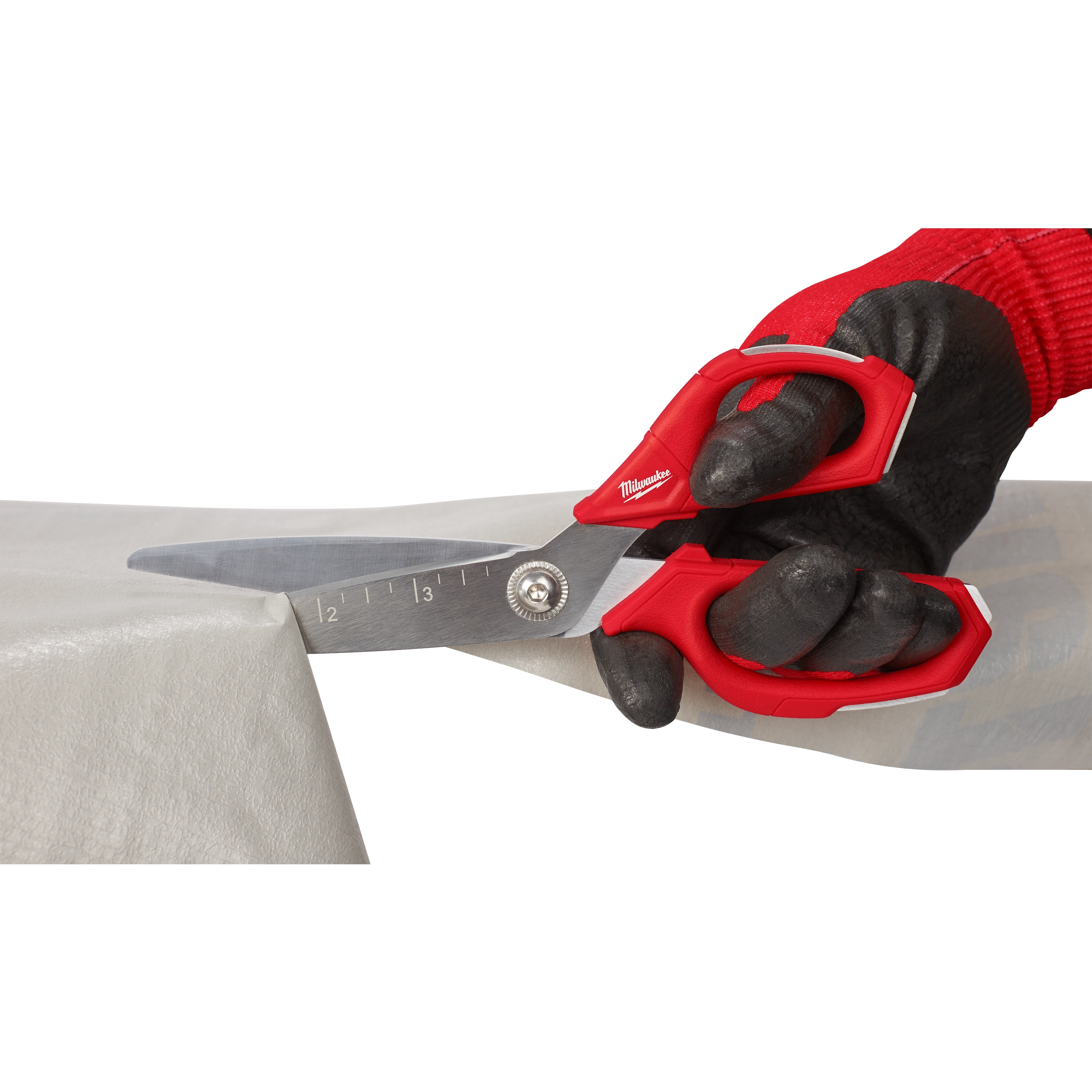 A gloved hand uses Offset Jobsite Scissors with a red handle to cut through a piece of thick, opaque material. The scissors feature measurement markings on the blade for precision.
