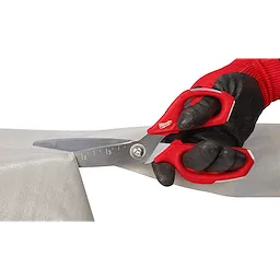 A gloved hand uses Offset Jobsite Scissors with a red handle to cut through a piece of thick, opaque material. The scissors feature measurement markings on the blade for precision.