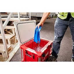 A person dips a blue Cooling Towel into a red cooler filled with water. The person wears a safety vest and jeans.