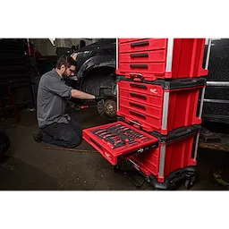 A mechanic utilizes tools from a 366-piece Master Mechanics Hand Tool Set with PACKOUT™ Drawers and Dolly. The red drawer system is stacked and open, showcasing the organized tools while the mechanic works on a vehicle in a garage.