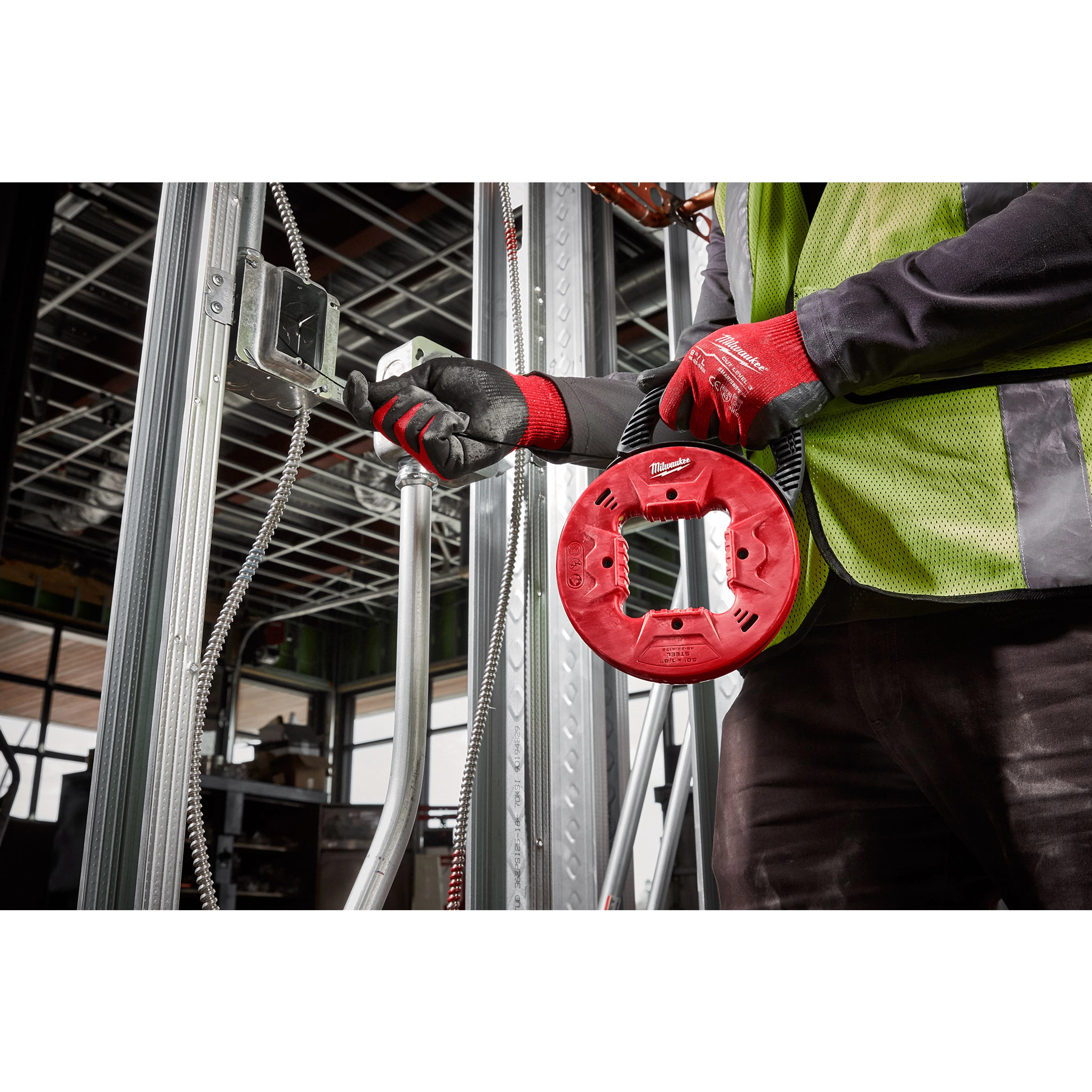 Worker using 1/8" 50' steel fish tape in a construction setting, wearing gloves and a safety vest, guiding tape through conduit.