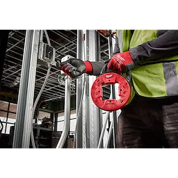 Worker using 1/8" 50' steel fish tape in a construction setting, wearing gloves and a safety vest, guiding tape through conduit.