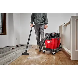 Person using the Milwaukee M18 FUEL 9 Gallon Dual-Battery Wet/Dry Vacuum to clean up wood debris on a wooden floor in a room.