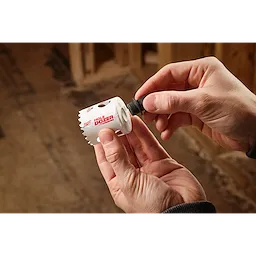 A person is holding a white Hole Dozer hole saw and attaching a 5PK Large Thread Adapter for QUIK-LOK™ Arbor. The background shows a partially constructed wooden wall.